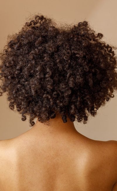 Back view of multiracial female model, standing with bare shoulders and demonstrating healthy, soft skin, posing against beige wall